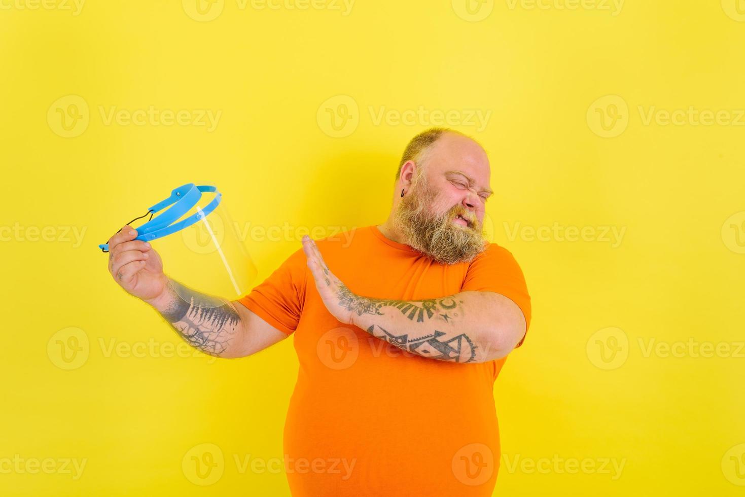 Doubter man with beard and tattoos is worried about a protective face shield against covid-19 photo