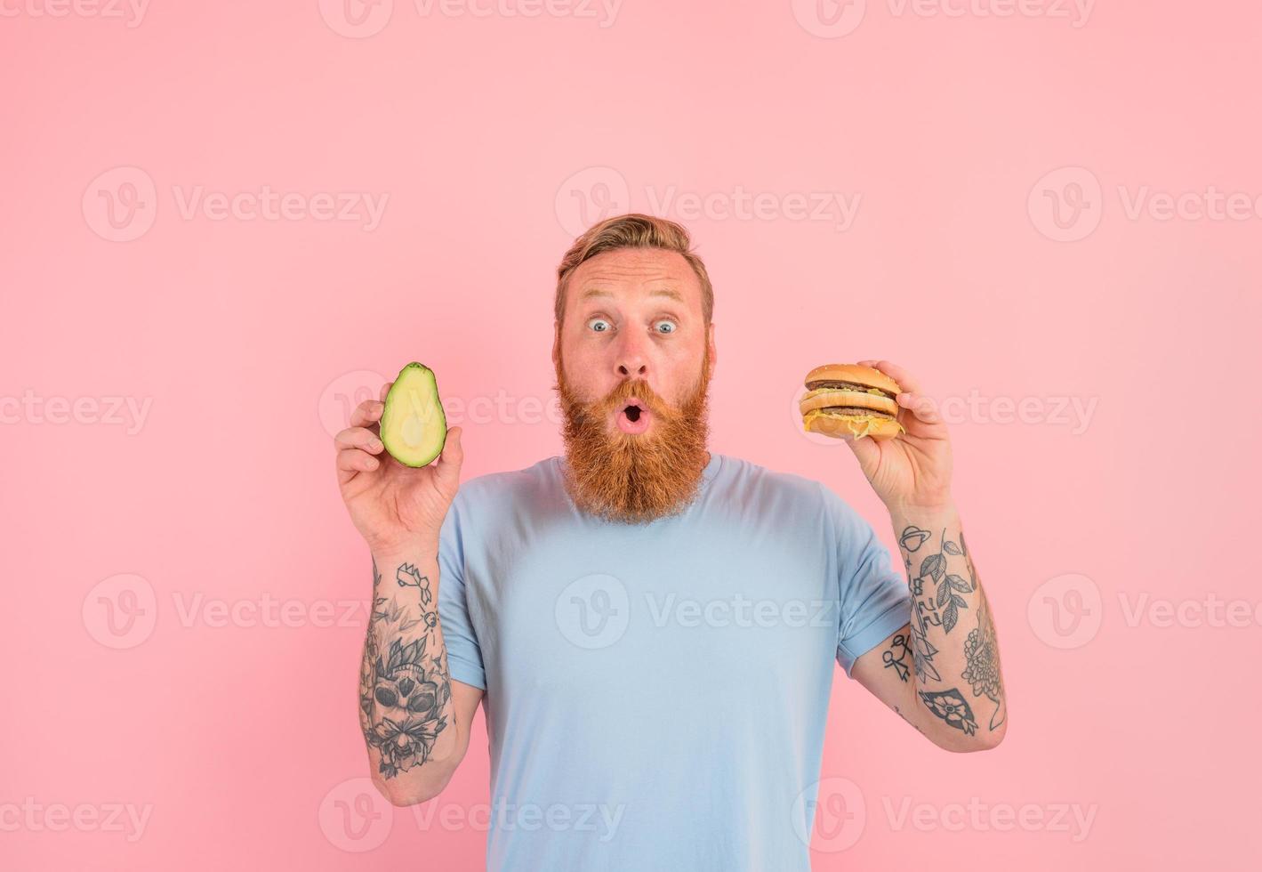 Amazed man with beard and tattoos is undecided if to eat an avocado or an hamburger photo