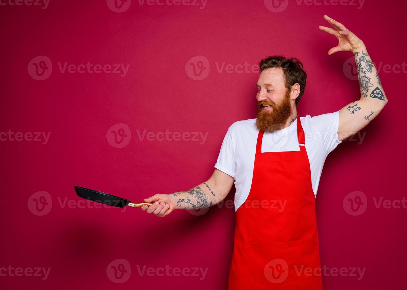 Happy chef with beard and red apron cooks with pan photo