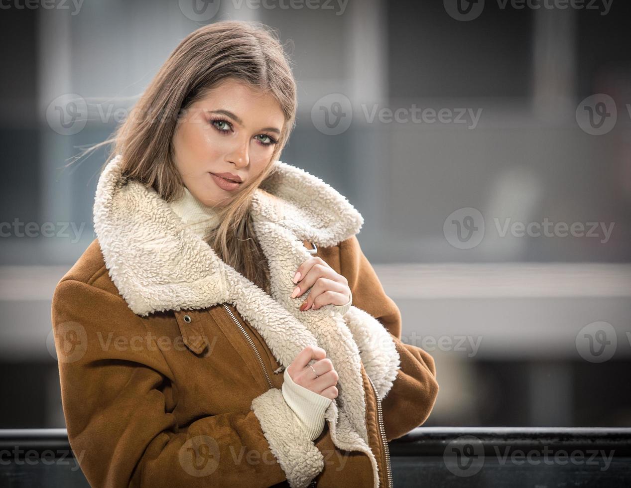 maravilloso sonriente niña en elegante ropa posando en moderno balcón con increíble ver en ciudad .retrato de un elegante de moda rubia Adolescente con piel Saco con grande collares vistiendo en balcón foto