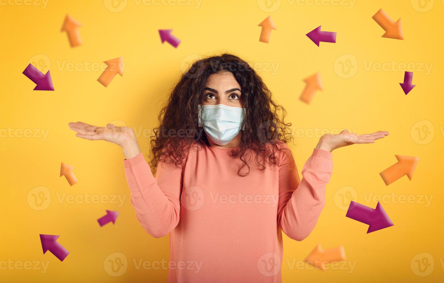 Girl with face mask is confused which arrow to follow. Concept of doubt about laws governing covid-19. Yellow background photo