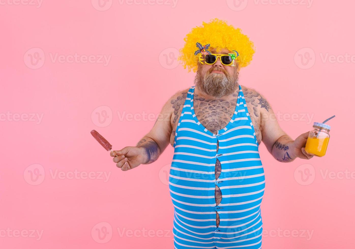 Fat confused surprised man with beard and wig eats a popsicle and drinks a juice fruit photo