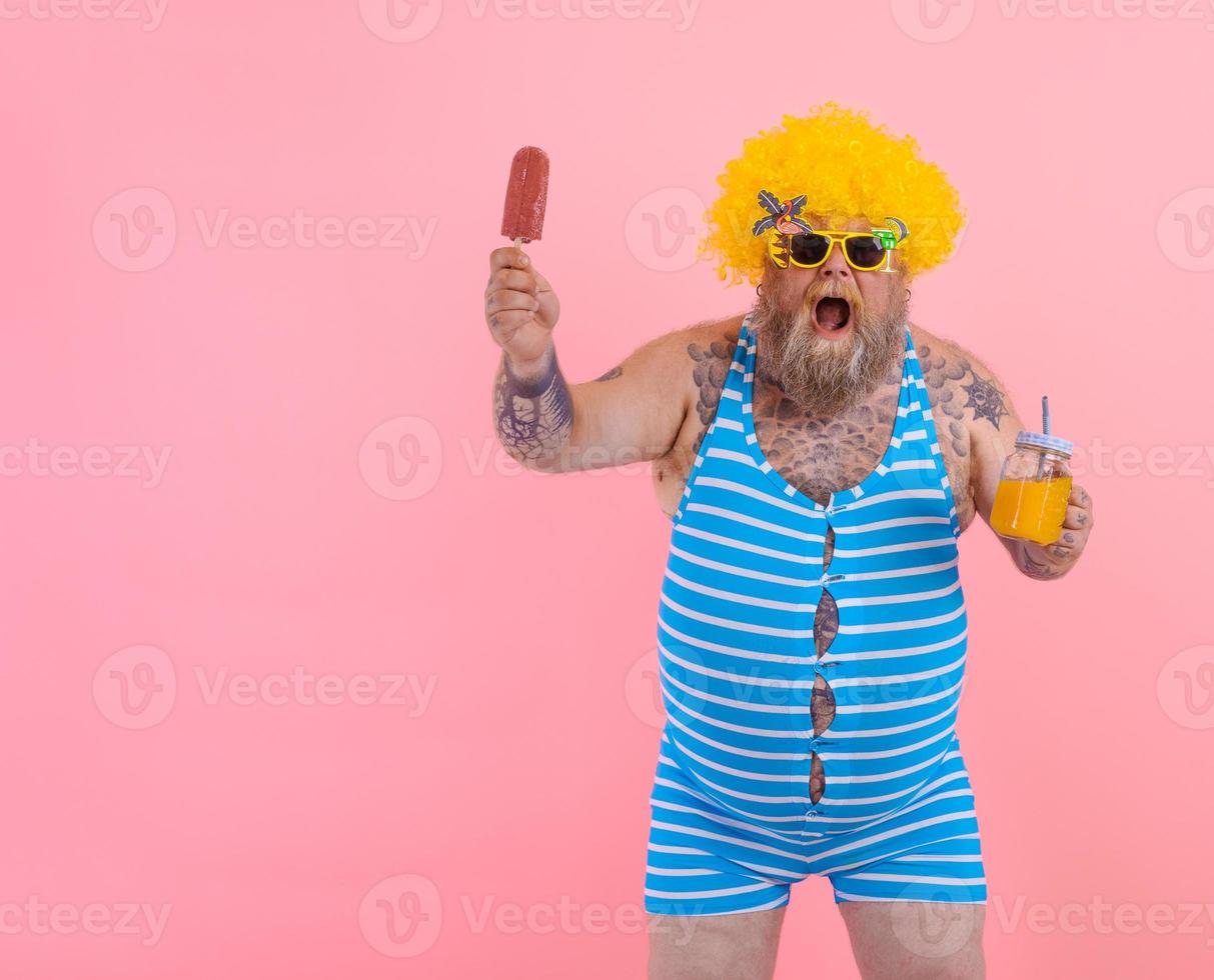 Fat surprised man with beard and wig eats a popsicle and drinks a juice fruit photo