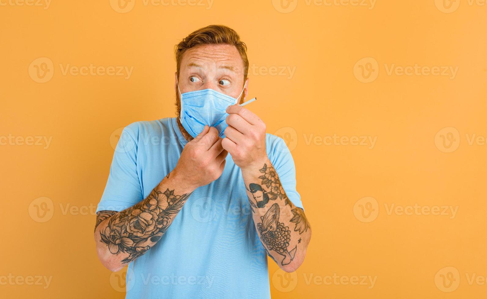 Afraid man with beard, tattoo and mask for covid-19 wants to smoke a cigarette photo