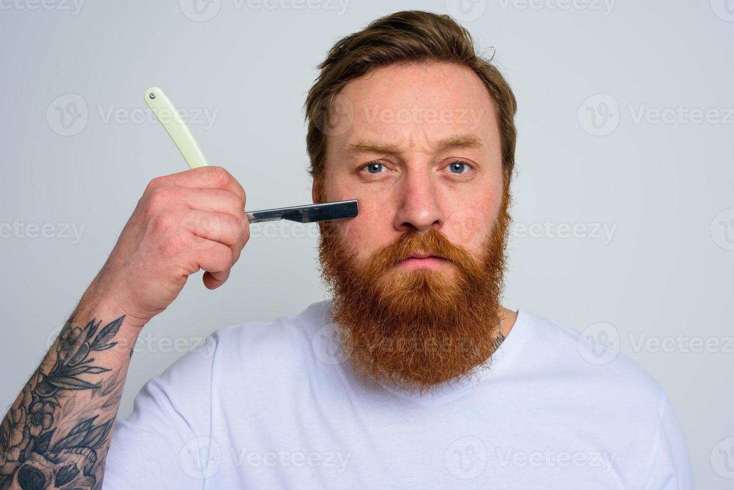 Serious man with blade is focused on cutting his beard photo