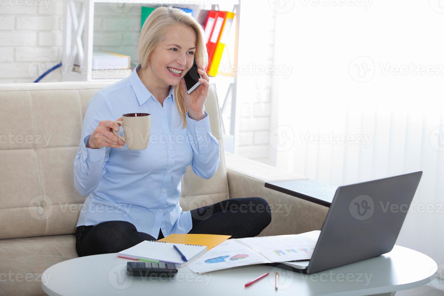 Listening to clients requirements. Beautiful middle aged woman with glasses talking on smart phone and smiling at her working place siting in the office. photo