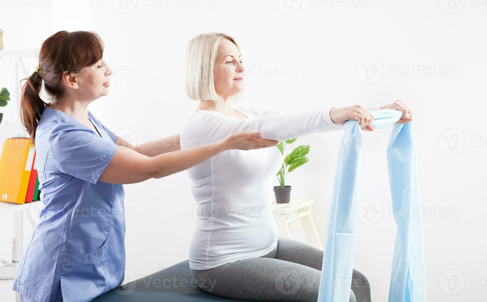 Physiotherapist and woman sitting on a bed exercising with a rubber tape photo