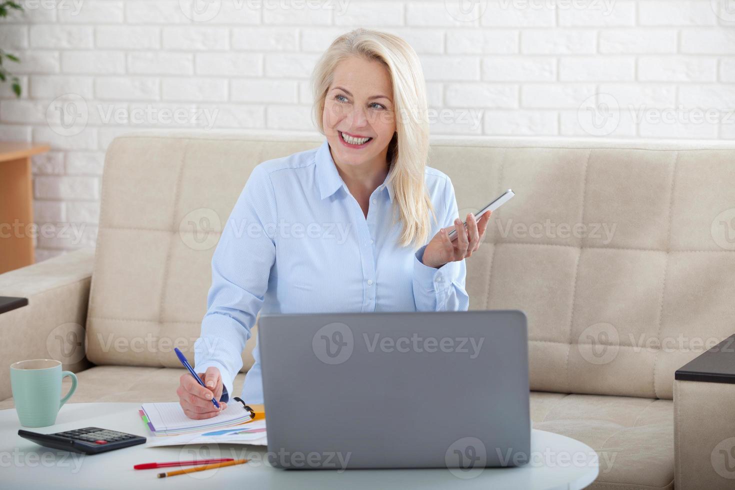 Listening to clients requirements. Beautiful middle aged woman with glasses talking on smart phone and smiling at her working place siting in the office. photo