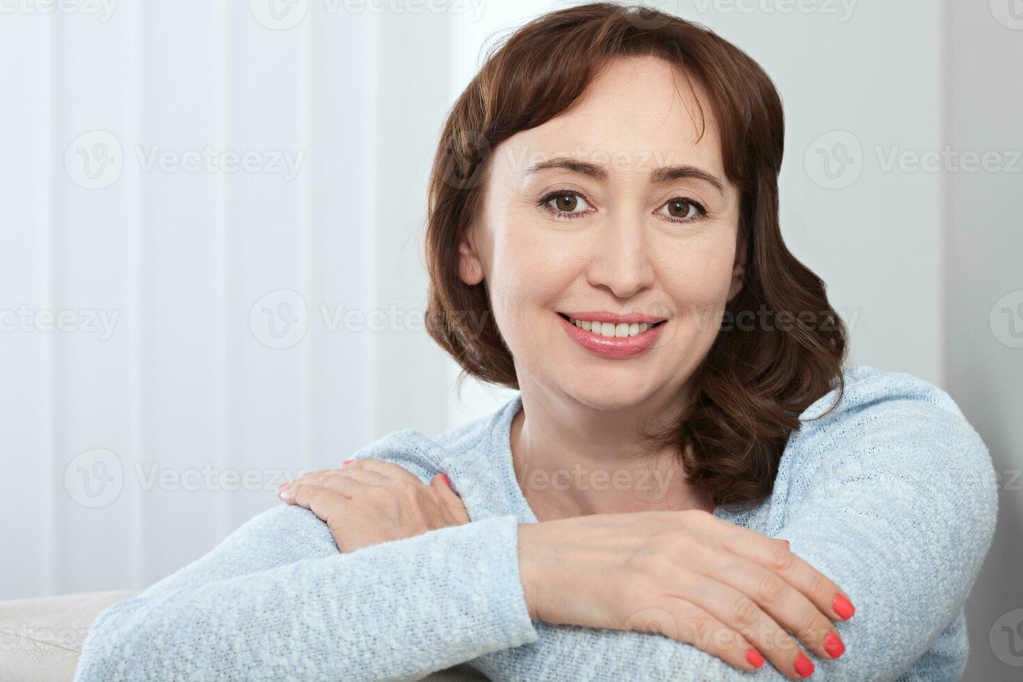 Lovely middle-aged brunette woman with a beaming smile sitting on a sofa at home looking at the camera photo