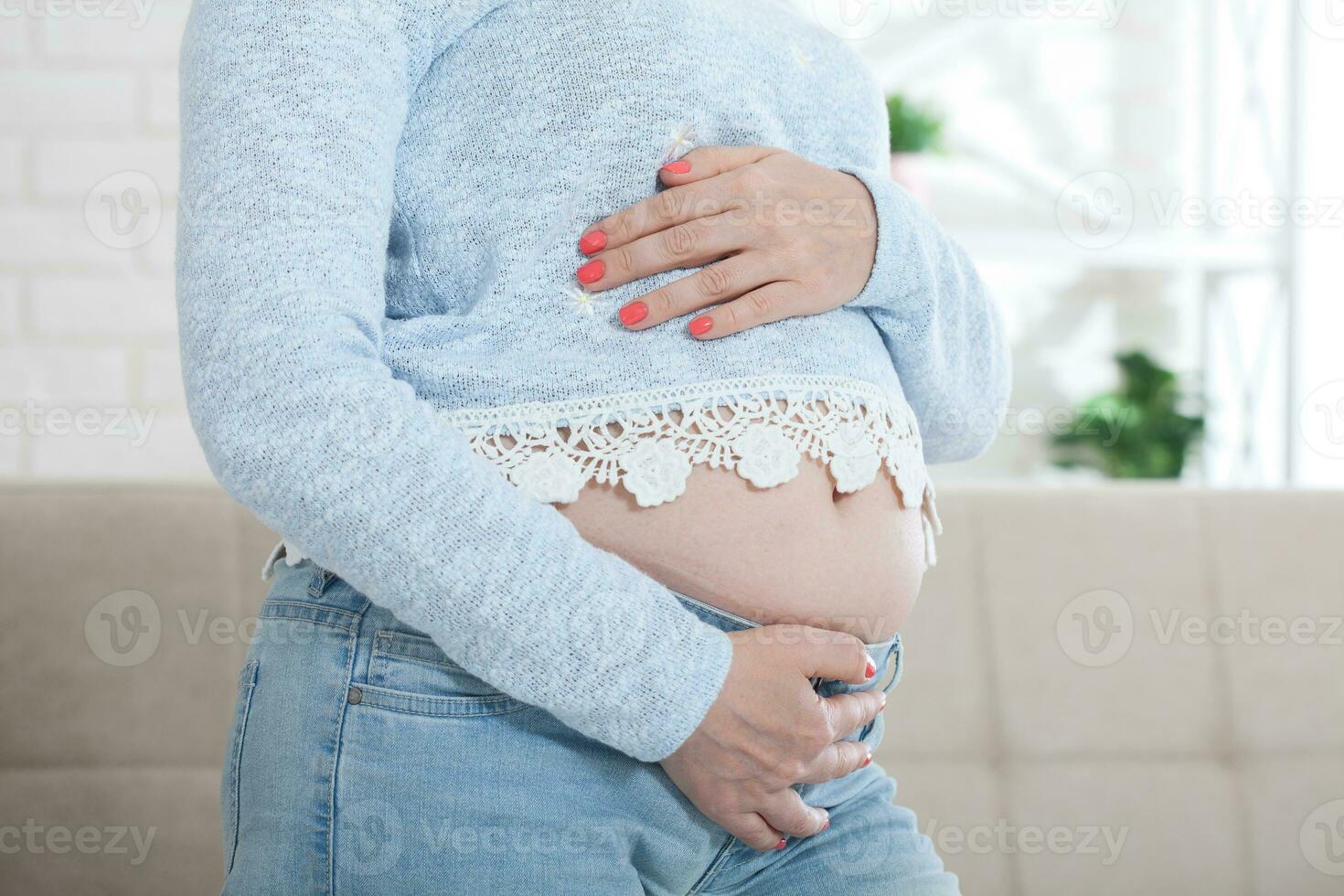 Pregnant middle aged woman in jeans holds hands on belly on white background. Pregnancy, maternity, preparation and expectation concept. photo