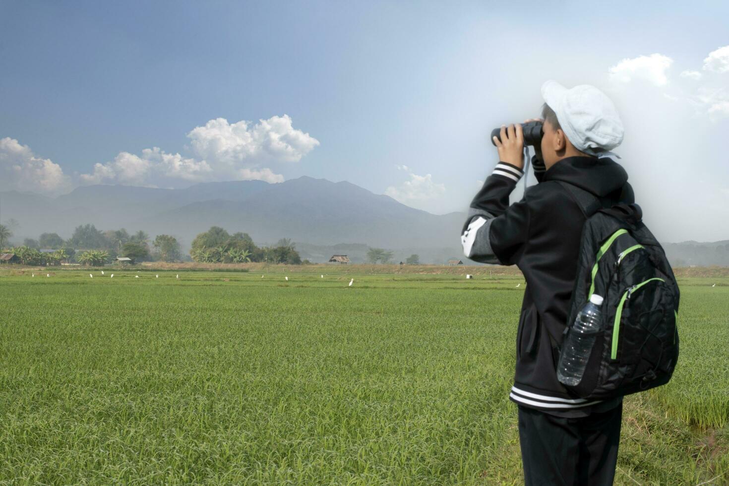 Asian boy in plaid shirt wears cap and has a backpack, holding a binoculars, standing on ridge rice field of asian farmers to observe the pm 2.5 smoke, dust and birds on tree branches and on sky. photo