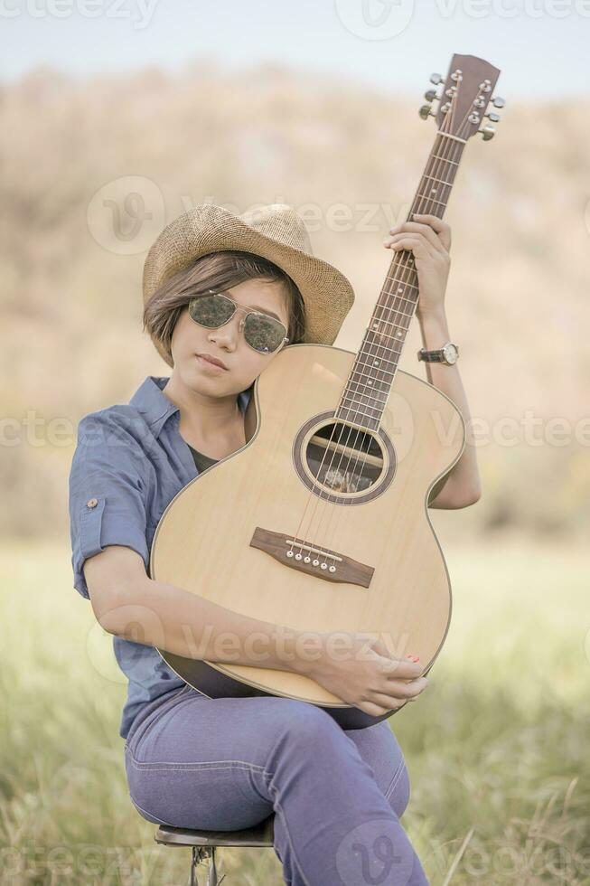 Women short hair wear hat and sunglasses sit playing guitar in grass field photo