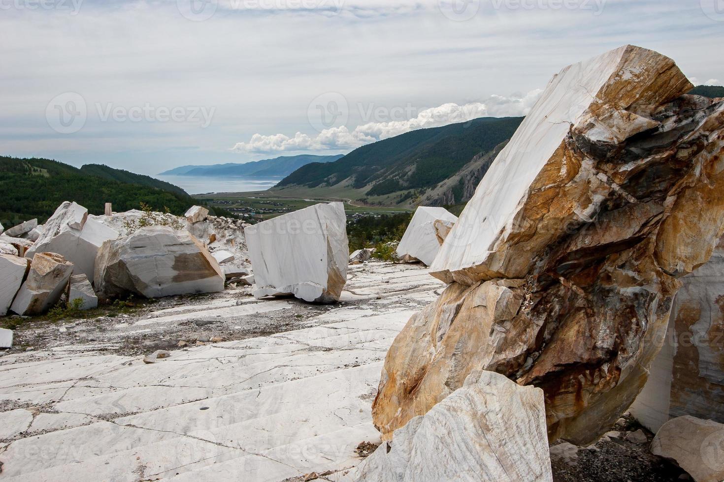 Large white marble blocks in an old abandoned quarry on the background of hills and Lake Baikal. Smooth cuts at the stones. Cloudy. Horizontal. photo