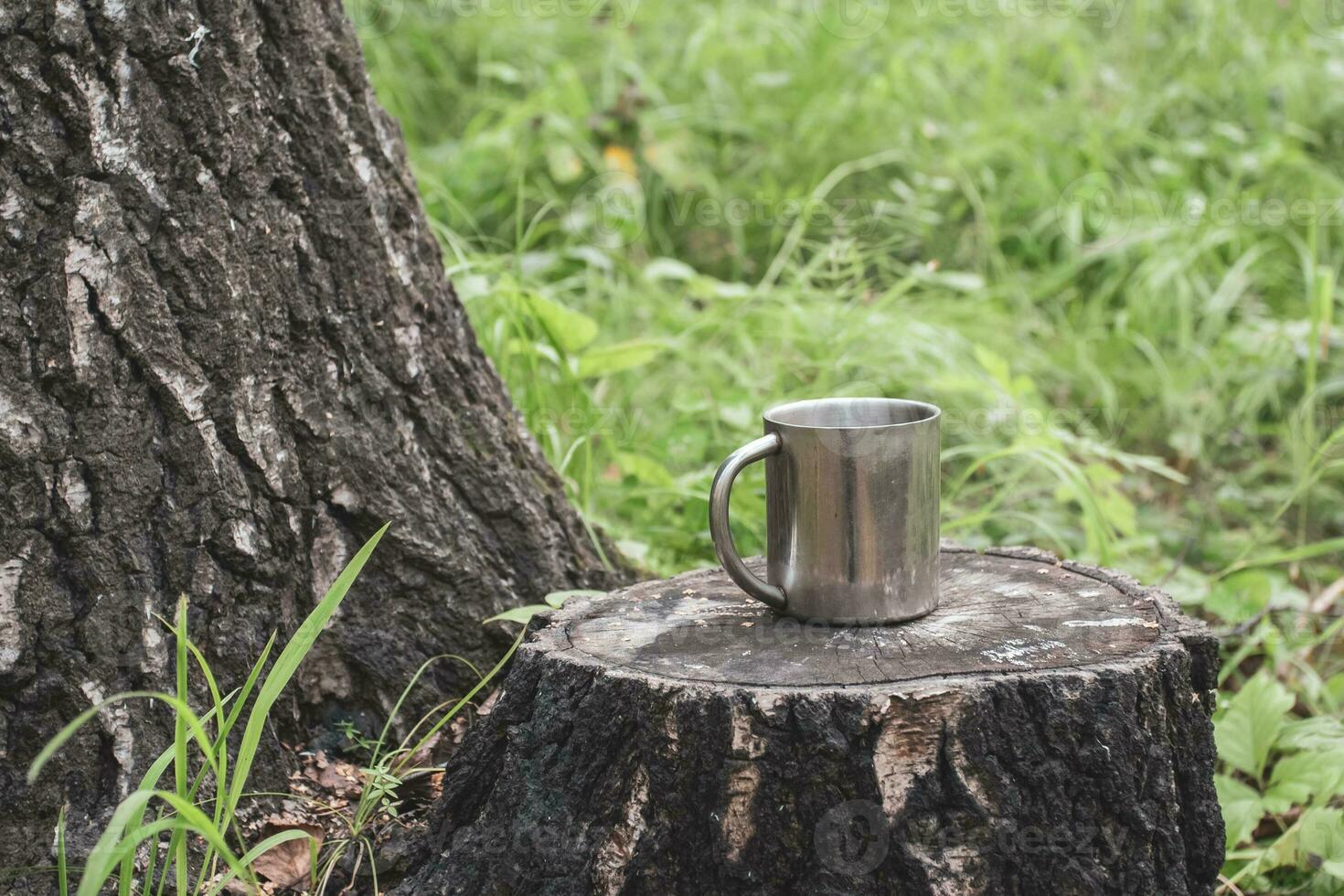 An iron tourist mug stands on a stump in a forest next to a thick tree against a background of blurred grass. Selective focus on the mug. The background is blurry. Place for text. photo