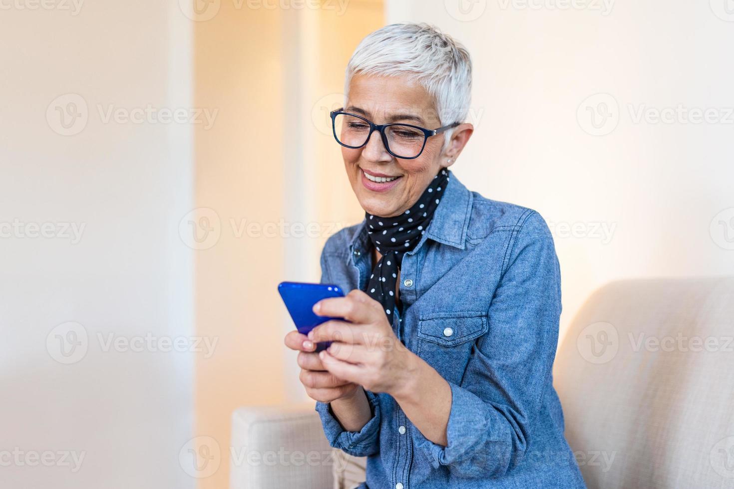 Happy senior woman using mobile phone at home. Smiling cool old woman with white hair sitting on sofa and messaging with smartphone. Beautiful stylish elderly lady talking on cellphone photo