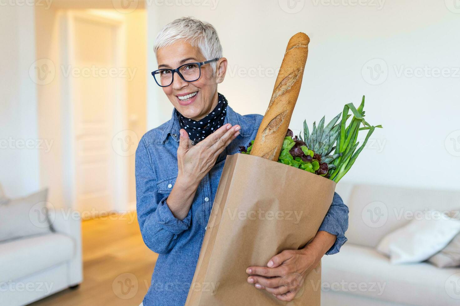 Happy mature woman holding a paper bag full of groceries from the supermarket. Elderly woman with groceries at home photo