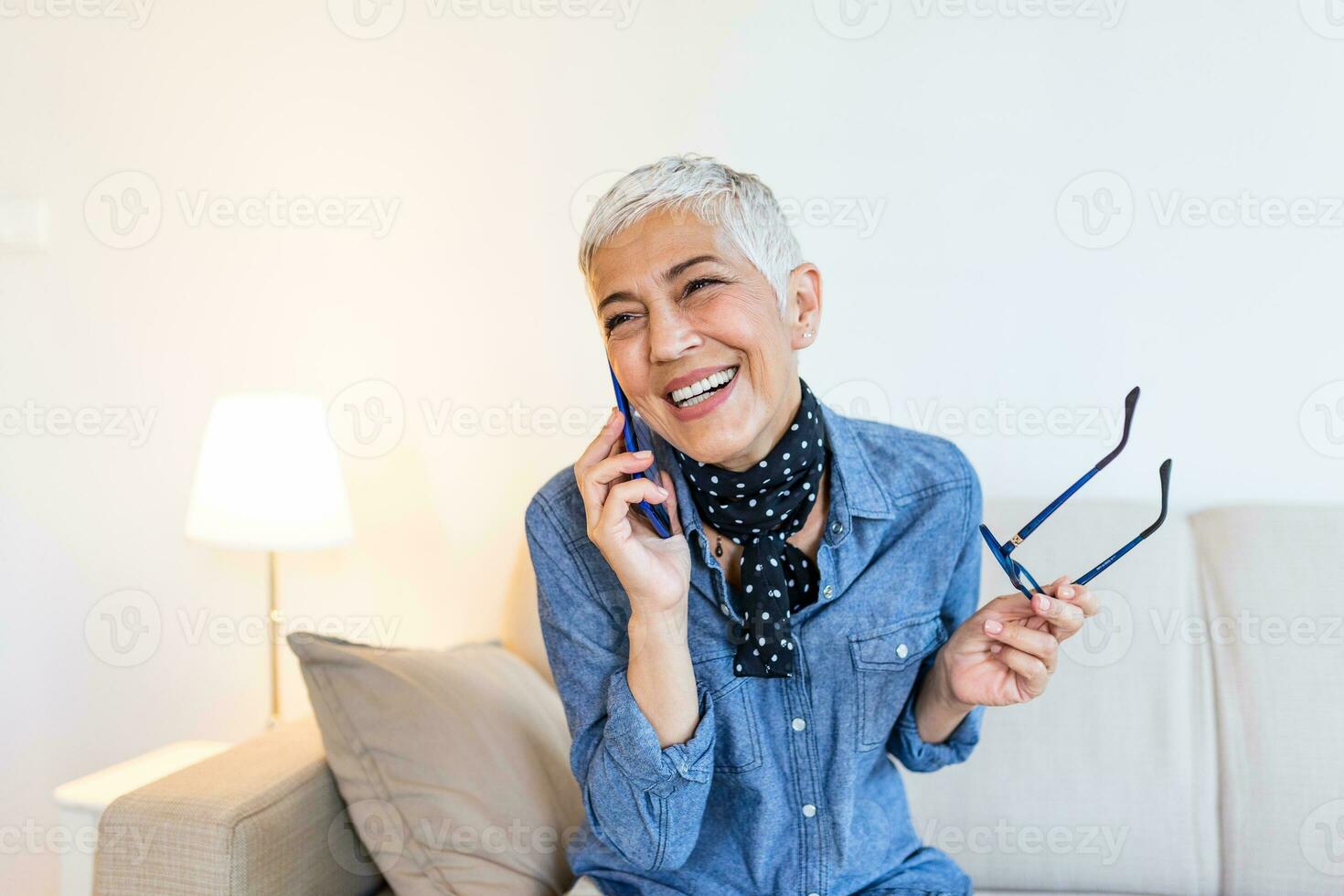 Happy senior woman using mobile phone at home. Smiling cool old woman with white hair sitting on sofa and messaging with smartphone. Beautiful stylish elderly lady talking on cellphone photo