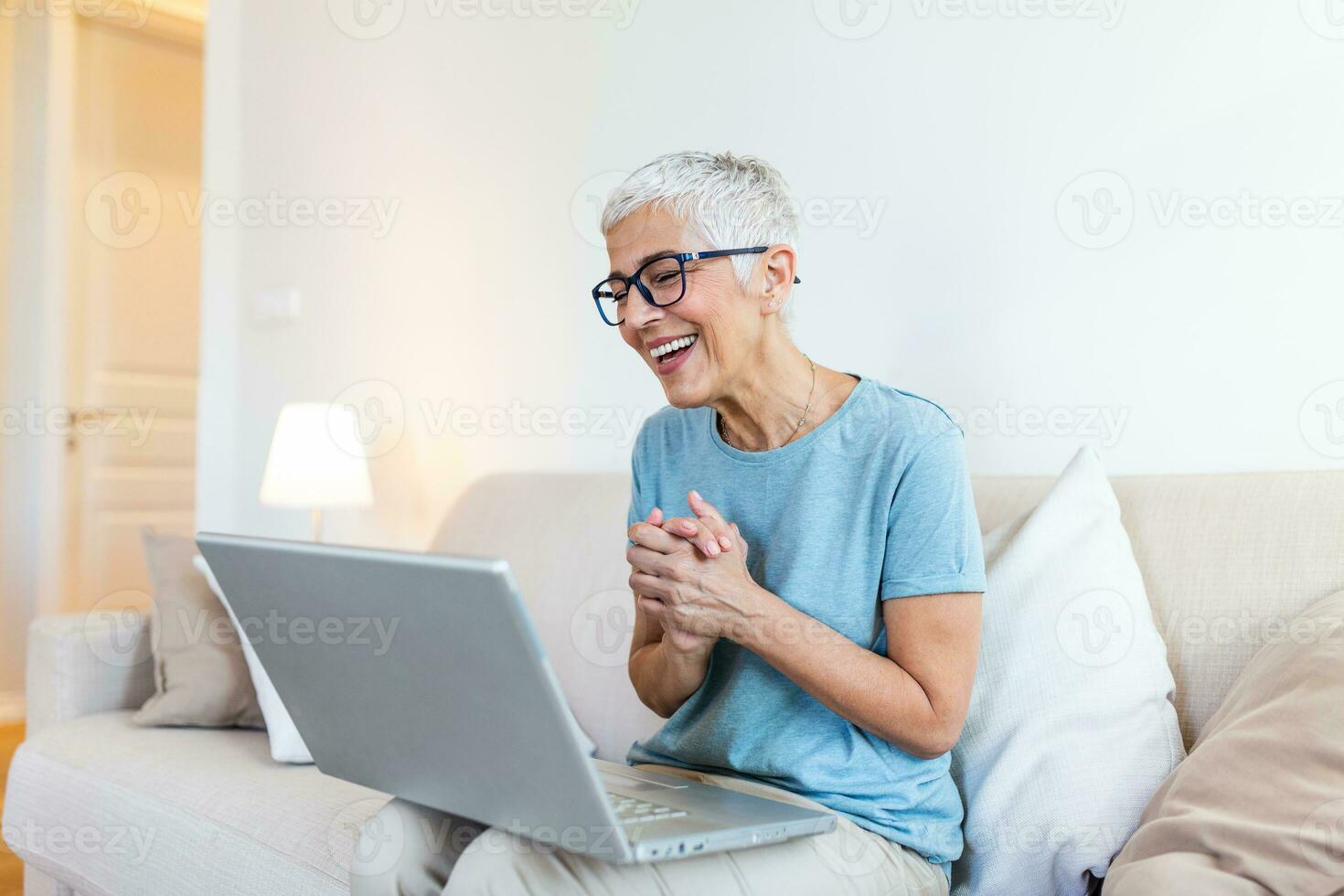 Happy mature woman waving to someone while having a video call over laptop at home. Gray-haired senior woman waving hand in front of laptop while having video call with her family members. photo