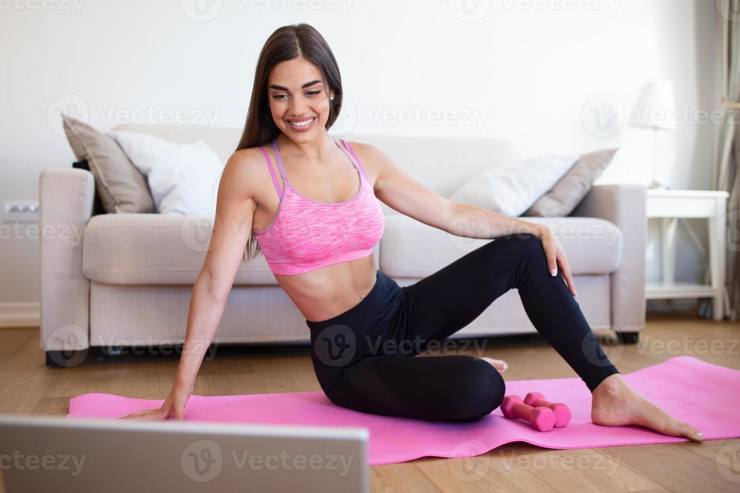 Gorgeous young woman on exercise mat at home and watching workout tutorial online on laptop. Video lesson. Young woman repeating exercises while watching online workout session photo
