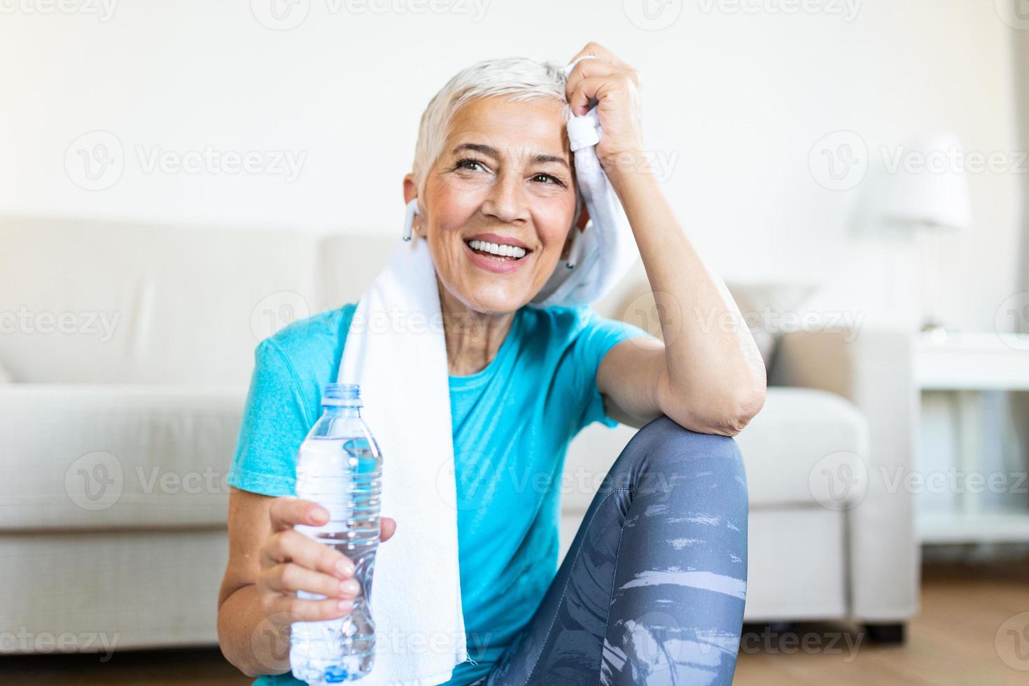 Senior woman holding plastic bottle of water,wiping sweat with a towel, exhausted after the daily training. Elderly woman taking a break while exercising at home. sitting on fitness mat and resting photo