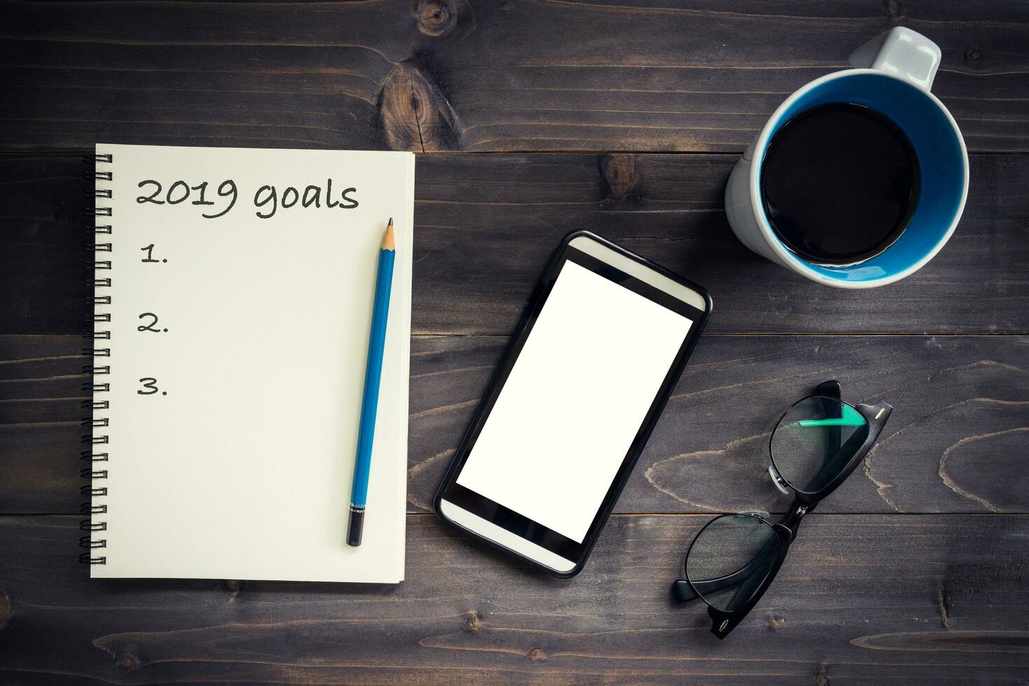 Notebook with 2019 goals massage with pencil, glasses, phone and cup of coffee on wood background. photo