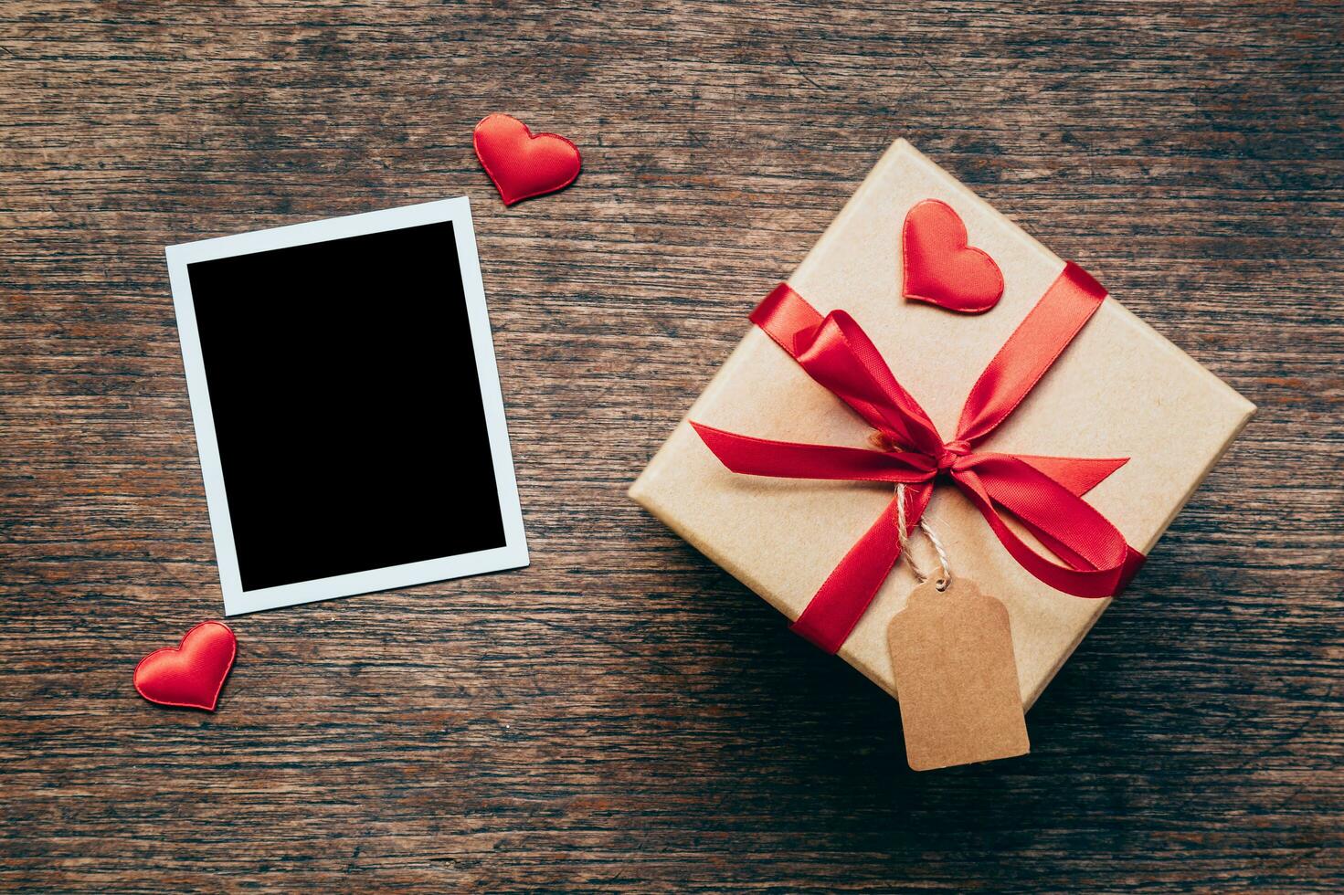 Blank photo frame and gift box with red heart on wood background.