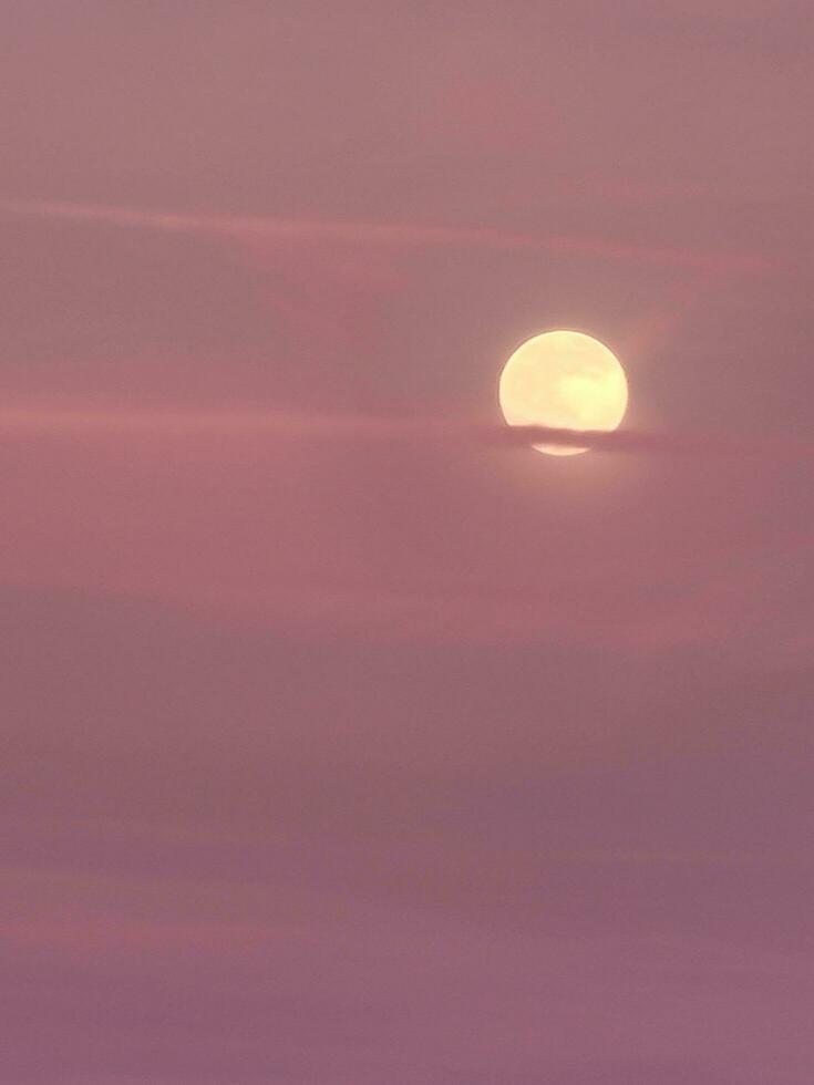 A hazy yellow moon peeking out through the pink and purple sunset sky photo