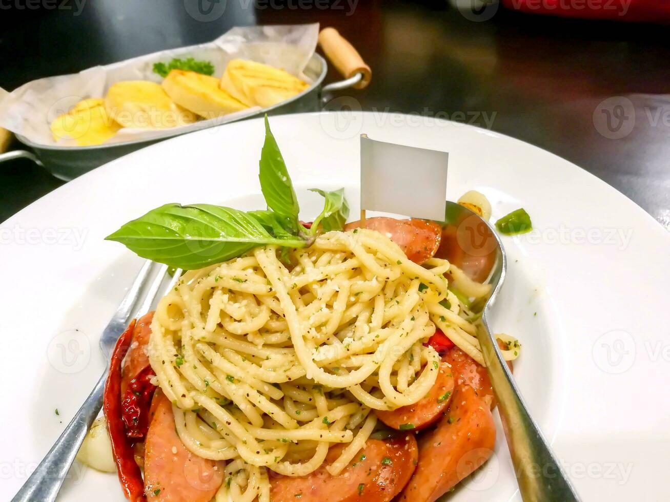 Sausage spaghetti in Thai style on white ceramic plate with spoon and fork and garlic bread in aluminium plate and wooden table in the restaurant. photo