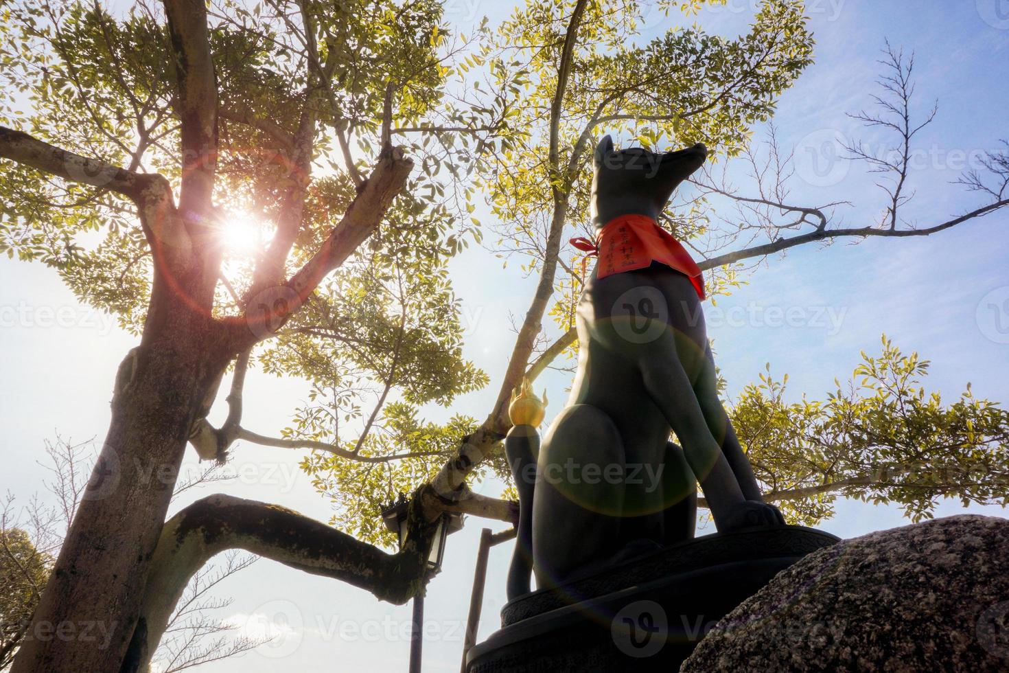 Fox statue of Fushimi Inari Shrine front of a big trees with sun and lens flare on blue sky background. photo