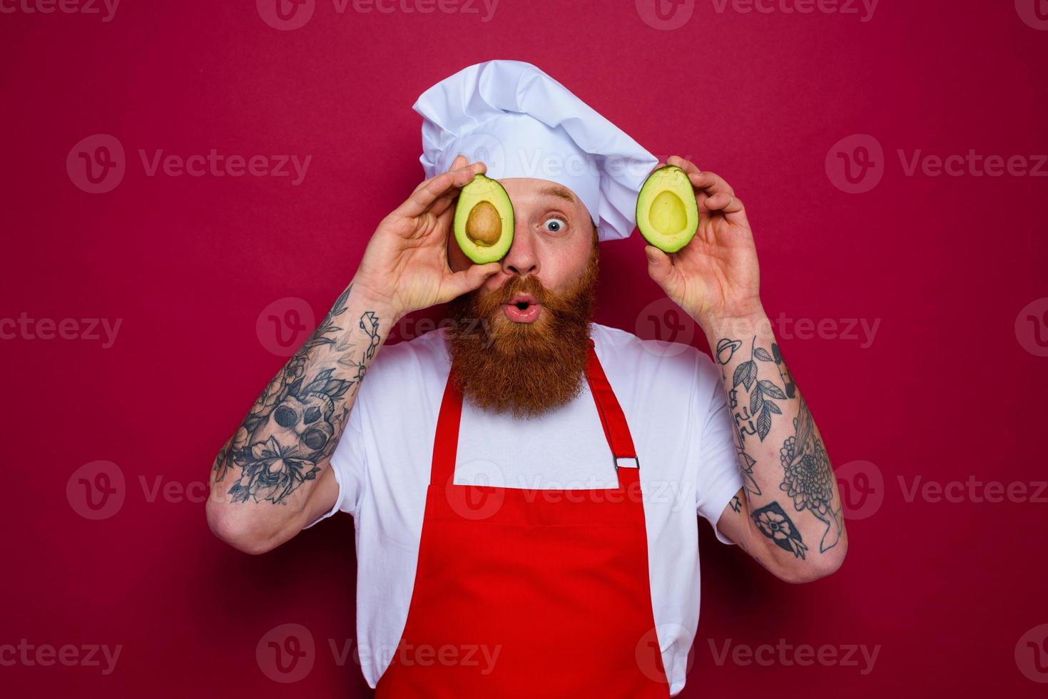 surprised chef with beard and red apron holds an avocado photo