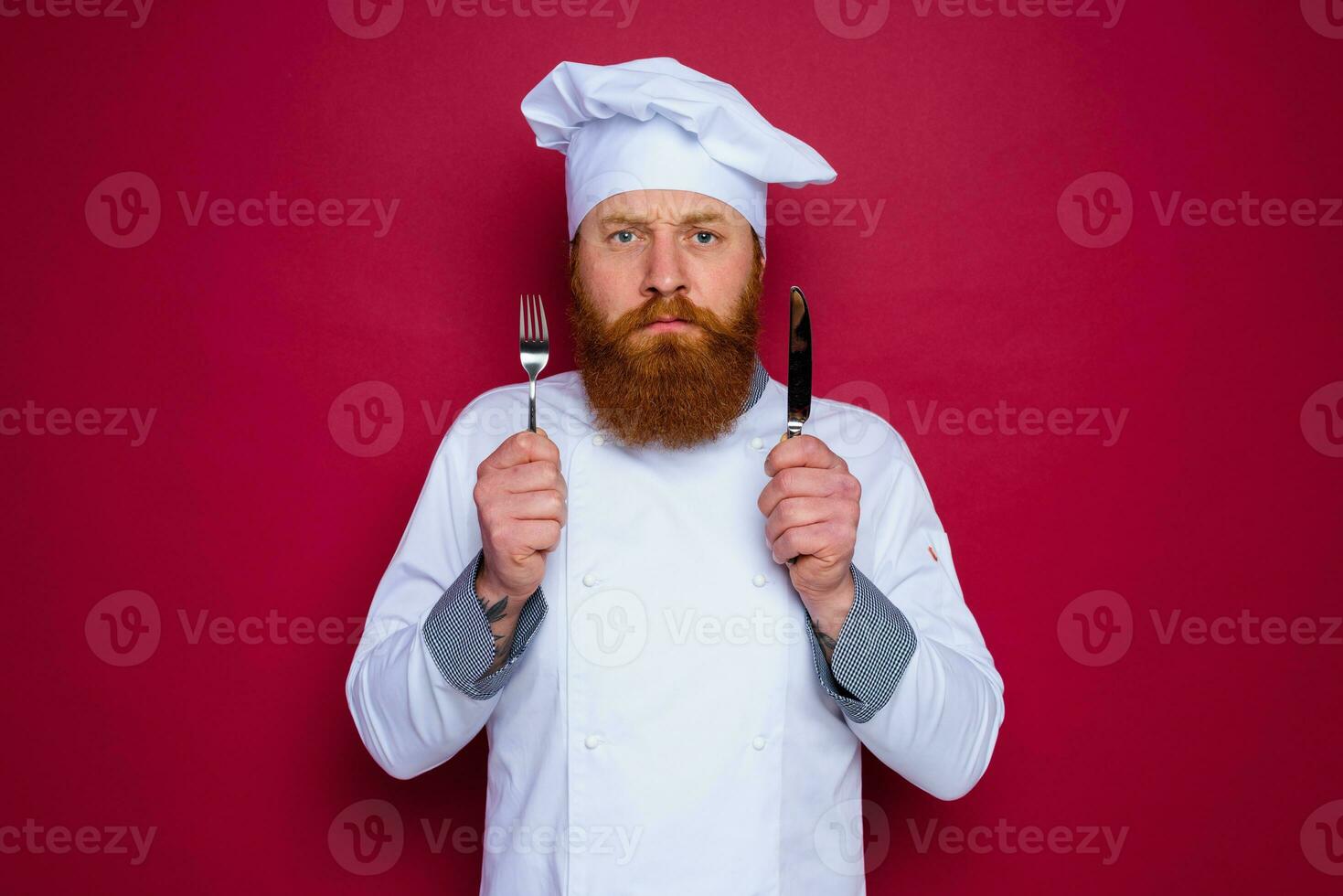 serious chef with beard and red apron holds cutlery in hand photo