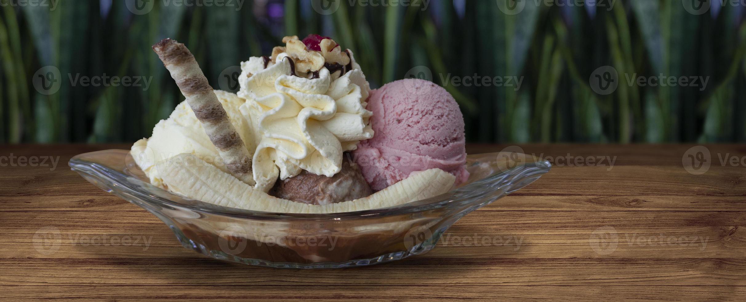 plátano división adornado con azotado crema y chocolate sorbete en claro vaso Pato en de madera mesa con desenfocado verde plantas antecedentes foto