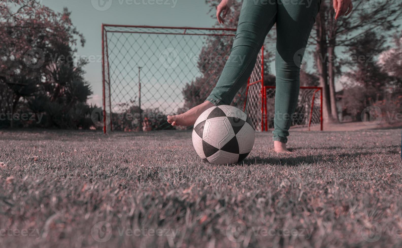 legs of woman in blue pants, barefoot in middle of park playing with a soccer ball with a soccer goal with nets and trees in background during the morning photo