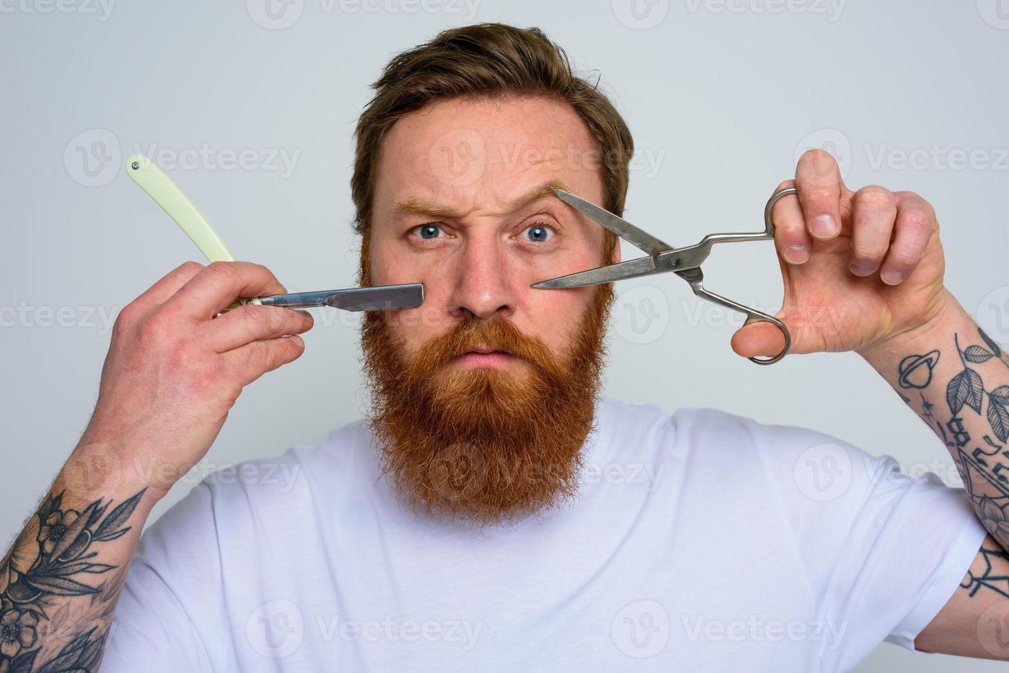 Worried man with scissors and blade is ready to cut the beard photo