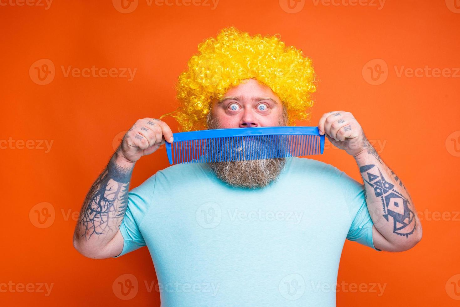 Fat amazed man with beard, tattoos and sunglasses combs himself with a giant comb photo