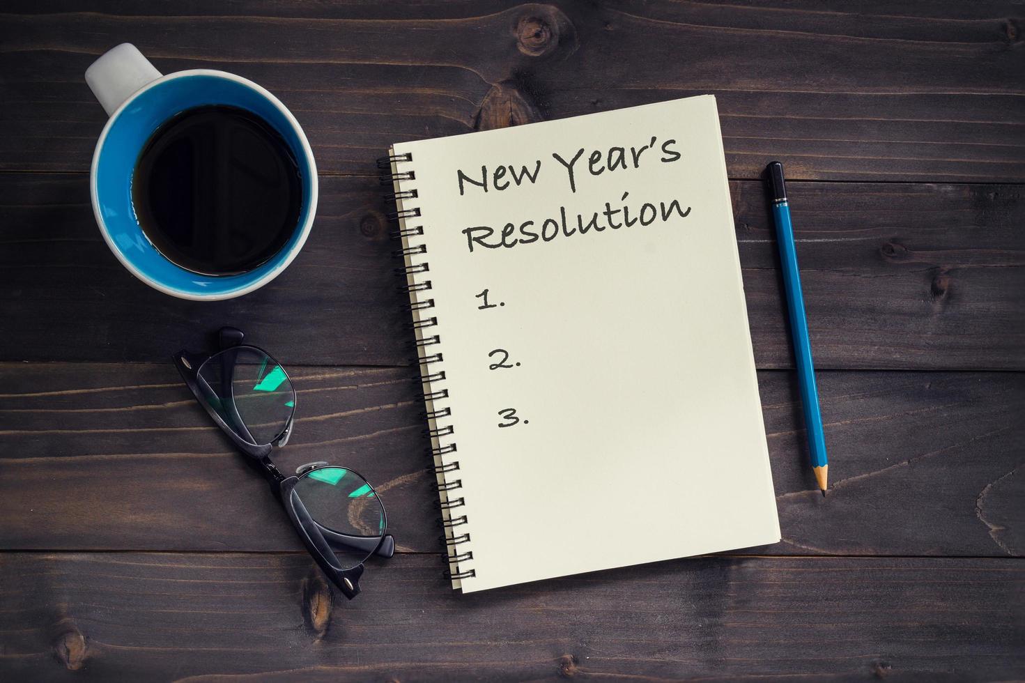 Notebook with New's Year Resolutions massage, pencil, glasses and cup of coffee on wood background. photo