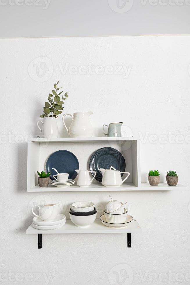 many ceramic and porcelain dishes stand on white open kitchen shelves with indoor flowers in pots. stylish interior view of part of the kitchen. photo