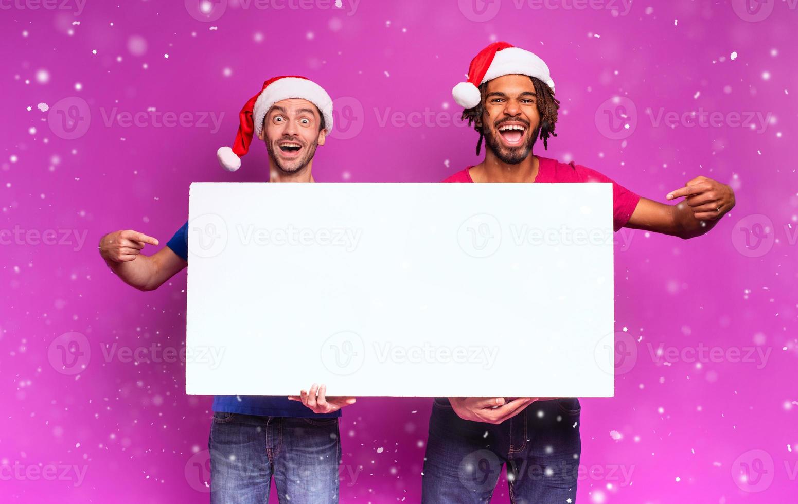 Surprised Amazed friends with Christmas hat hold a white banner for your message. Purple background photo