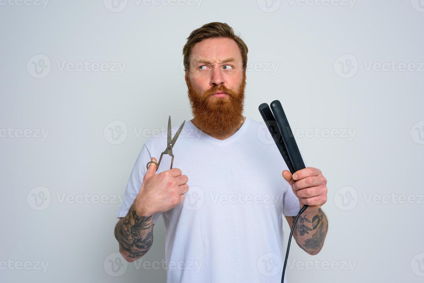 Undecided man with scissors and straightener is ready to work with hair photo
