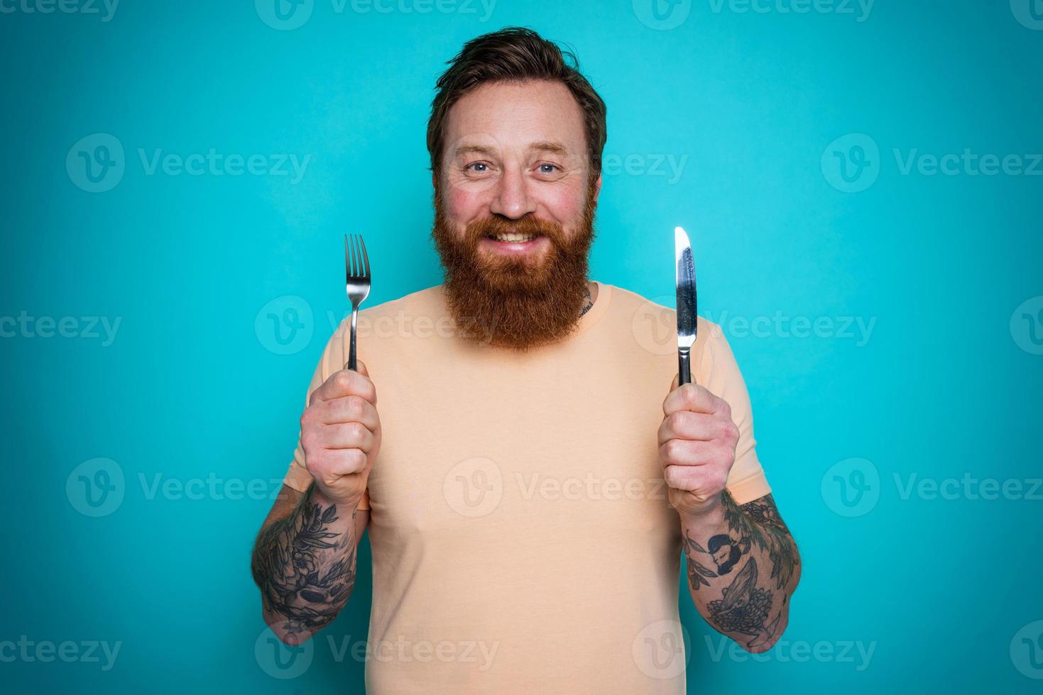 Man with tattoos is ready to eat with cutlery in hand. cyan background photo