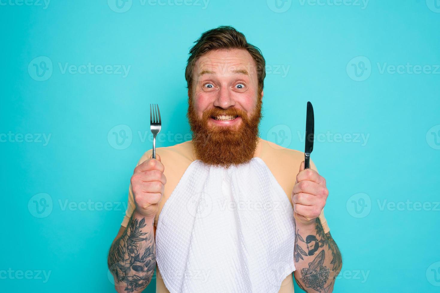 Happy man with tattoos is ready to eat with cutlery in hand photo