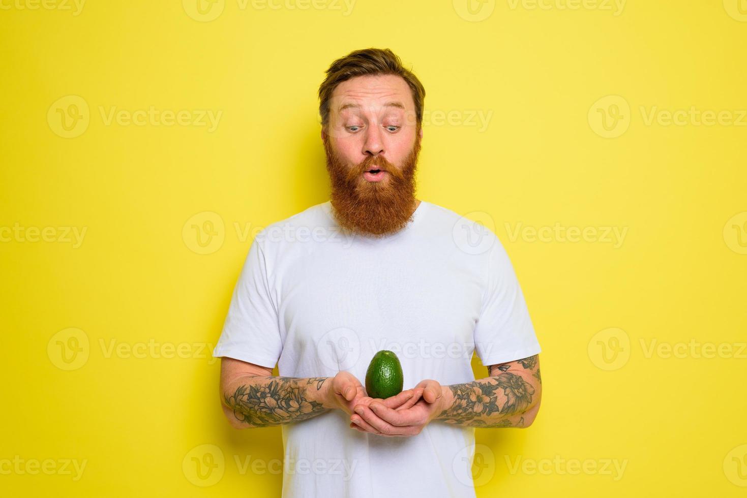Amazed with beard and tattoos holds an avocado photo