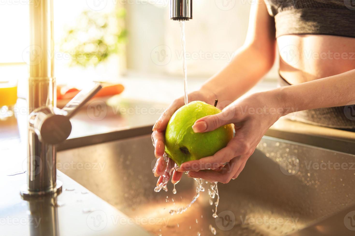 Young woman washes, with running water, an apple in the kitchen sink illuminated by the sun photo