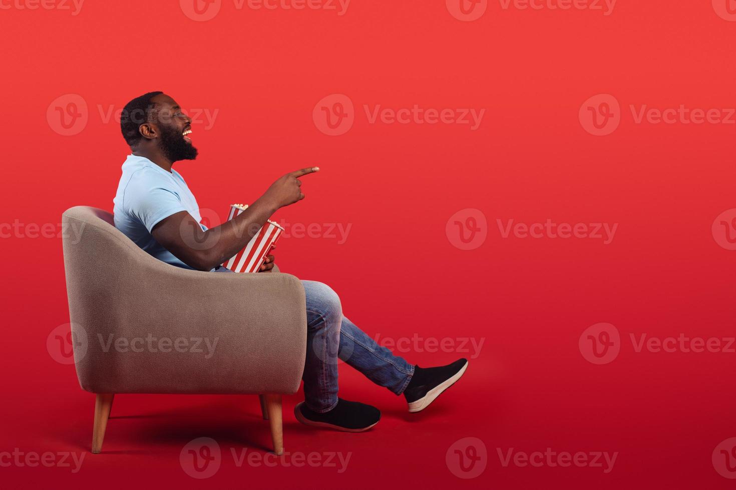 Amused televiewer sitting in the armchair at home while eating popcorns and watching a movie on red background photo