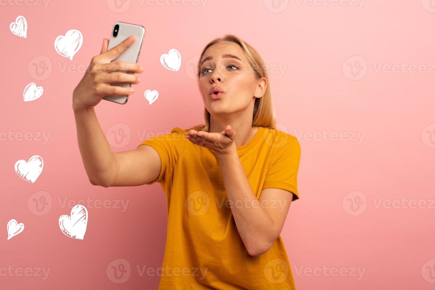 Blonde cute girl sends hearts on her smartphone. Happy and lovely expression face. Pink background photo