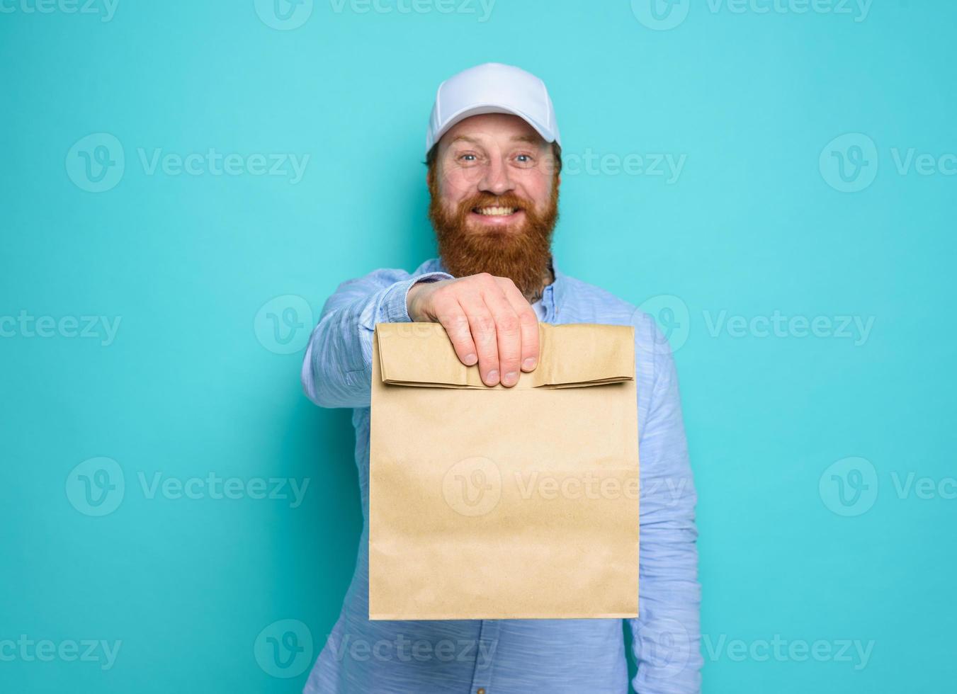 Deliveryman with wondered expression ready to deliver bag with food. cyan background. photo