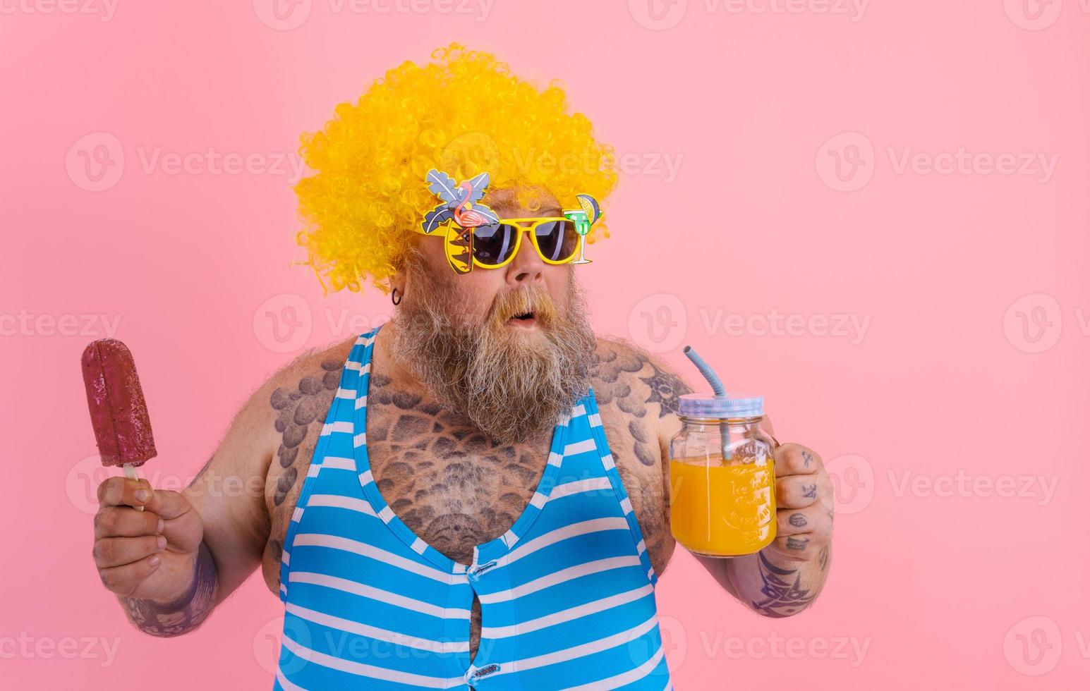 Fat pensive man with beard and wig eats a popsicle and drinks a juice fruit photo