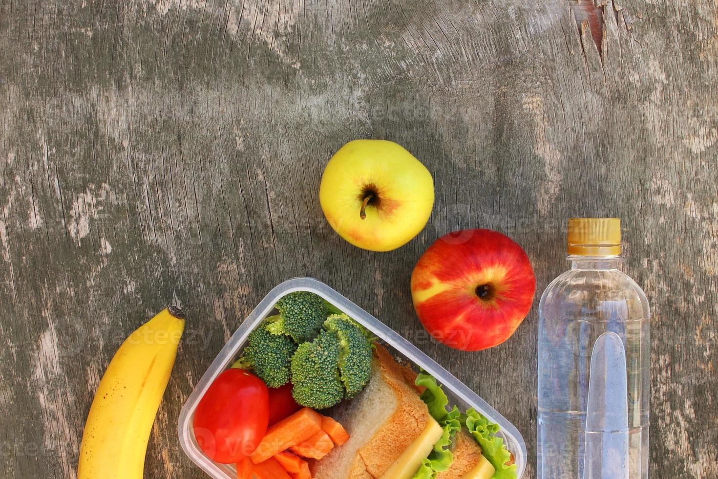 Sandwiches, fruits and vegetables in food box, water on old wooden background. Top view. Flat lay. photo