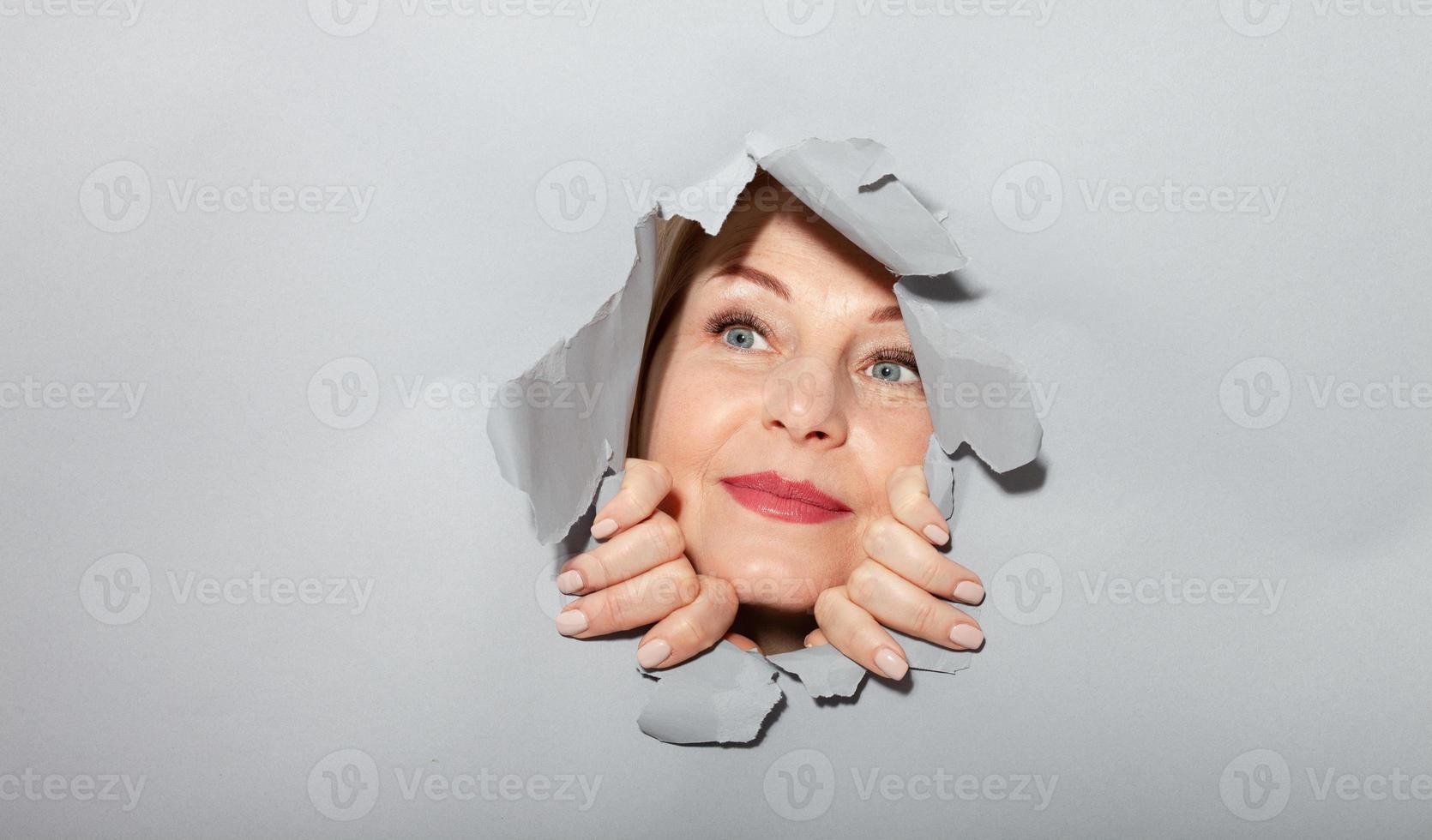 Surprised woman looking playfully in torn paper hole, has excited cheerful expression, looks through breakthrough of gray background. Wow, great news photo