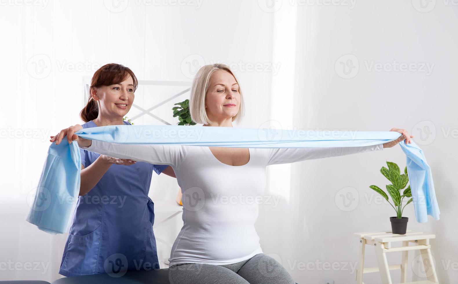 Physiotherapist and woman sitting on a bed exercising with a rubber tape photo